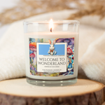 Glittering white soy wax candle titled "Welcome to Wonderland", inspired by the timeless adventure of Alice through the fantastical land of Wonderland. Candle sits lit atop a sliced tree trunk wooden platform with white cashmere blanket resting softly behind. Candle label features the White Rabbit with gold pocket watch, red bowtie, and blue blazer in center with a watercolor style mushroom forest in the background, Alice slowly making her way along its path in the bottom right corner.