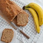 Banana Bread Soy Wax Candle Tin by Fervor Candle Company scent inspiration photo. White kitchen counter with grey and white checkered cotton napkin displaying fresh baked banana bread loaf and slices on a wooden cutting board with wooden-handled knife next to a bunch of perfectly ripe bananas.