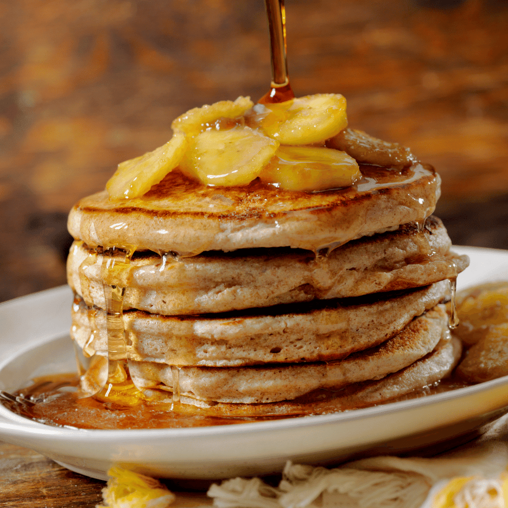 Banana Pancakes Hydrating Liquid Hand Soap by Fervor Candle Company scent inspiration photo. Pictured is a deep white porcelain platter with a tall stack of thick fresh pancakes, topped with sliced bananas and hot maple syrup.