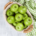 Fervor Candle Company Juicy Green Apple Room & Linen Mist scent inspiration photo. A white-washed wooden table with green and white checkered cloth displaying a linen-lined wicker basket full of shiny green apples.