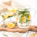 Fervor Candle Company Lemon & Rosemary Hydrating Liquid Hand Soap scent inspiration photo. A worn white-washed wooden farmhouse table under a wooden cutting board displaying a glass pitcher of water filled with lemon slices and vibrant green rosemary sprigs.