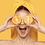 Orange Glow Hydrating Face Wash lifestyle image by Fervor Candle Company. Smiling woman wearing an orange towel hair wrap and holding sliced orange halves over her eyes.