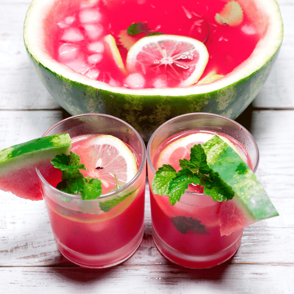 Fervor Candle Company Watermelon Lemonade Room & Linen Mist scent inspiration photo. A white-washed wooden farmhouse tabletop displaying a carved out watermelon half filled with hot pink watermelon lemonade, ice, and lemon slices, set behind two clear highball drinking glasses filled with the same hot pink watermelon lemonade, garnished with watermelon slices, lemon slice, and mint leaves.